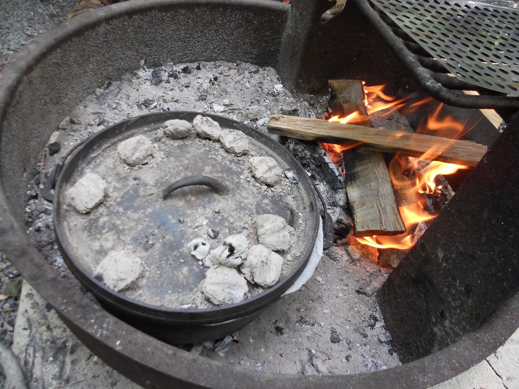 A cast iron Dutch oven with hot coals on the lid, cooking over a campfire