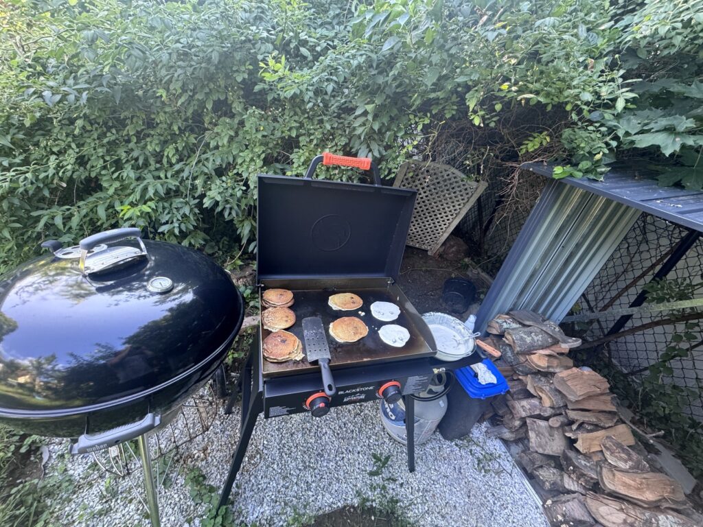 The Blackstone Adventure Ready 22-inch griddle on its stand, cooking pancakes on both burners in a backyard next to a charcoal grill